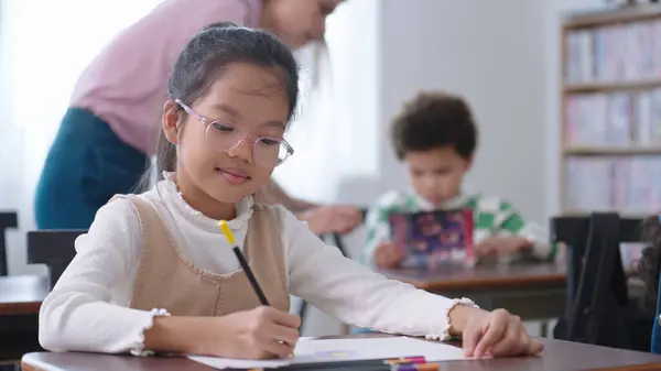 メガネをかけたアジアの女子生徒が教室でノートを書いている 女子高生は試験の準備と勉強をしています 小さな生徒が教室の机で書いている 教育知識コンセプト — ストック写真