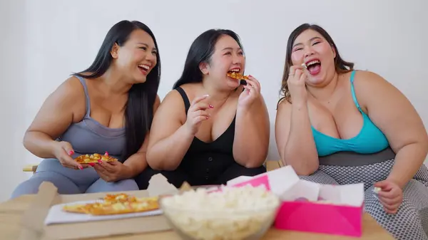 Group of Asian overweight woman friends enjoy eating unhealthy food whlie watching television together at home. Junk food, Fast food, Unhealthy food concept