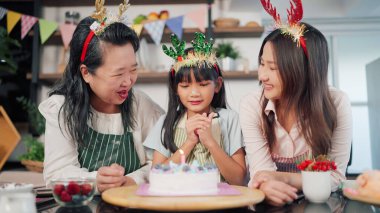 Asyalı küçük kız bir dilek diliyor ve anneanne ve anneyle birlikte evdeki mutfak odasında pastaya mum üflüyor. Evde aile kutlaması. Neşeli küçük kız ailesiyle birlikte doğum günü partisini kutluyor.