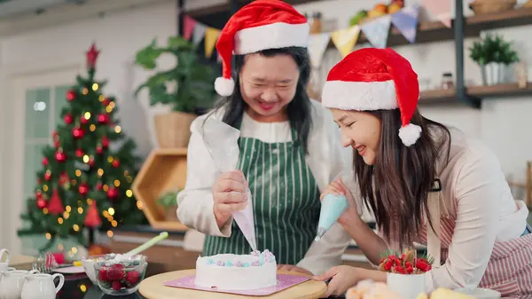 Happy asian elderly mother and young daughter helping squeezes cream onto a cake together. Mother and daughter making cake for Christmas day. Christmas cake decorated. Festive holiday