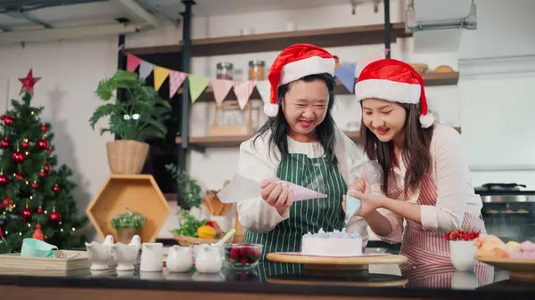 Happy asian elderly mother and young daughter helping squeezes cream onto a cake together. Mother and daughter making cake for Christmas day. Christmas cake decorated. Festive holiday