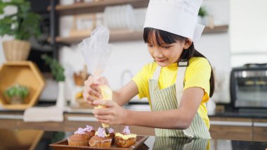 Önlüklü küçük Asyalı kız çocuğu ve aşçı şapkalı krem şanti ev yapımı kekleri ev mutfağında hazırlıyor. Ev yapımı kek hazırlayan ve dekore eden küçük bir kız. Çocuklar yemek yapıyor