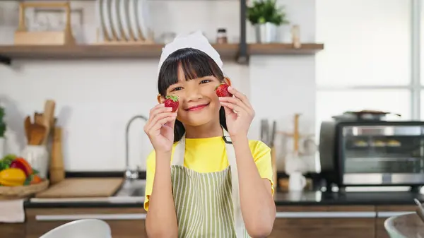 イチゴの笑顔とカメラを見ることによって目を閉じるエプロンのポートレート小さな幼い幼いアジアの少女 陽気な小さな女の子は楽しんでいる 手でイチゴを握る 目を覆う — ストック写真