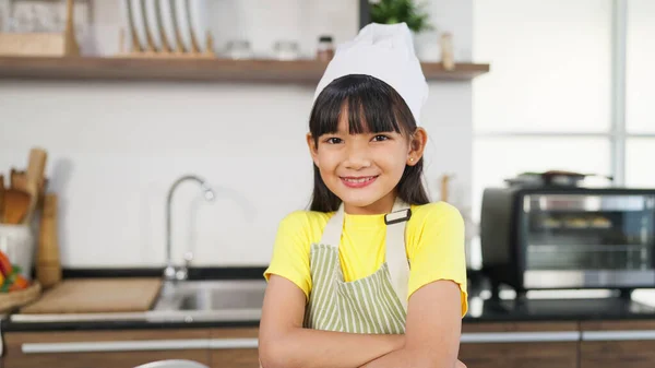 Portrait of asian little girl in apron standing with arms crossed and looking camera in kitchen at home. Cheerful little girl practicing to be a chef. Kid chef. Concept for cooking learning for kid