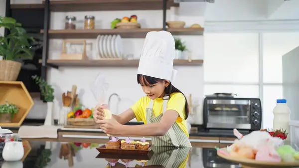 エプロンとシェフ帽子の小さな幼い幼い幼いアジアの少女は 家庭のキッチンで自家製のカップケーキを準備するクリームをホイップしました 小さな女の子が自家製のケーキを準備して飾ります 子供たちが料理する — ストック写真