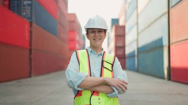 Güzel beyaz kadın ustabaşının gülümseyen portresi. Kasketli ve güvenlik yelekli. Kolları çapraz durarak kameraya bakıyor. Konteynır terminalinde müfettiş veya denetleyici