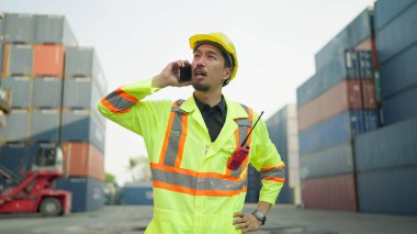 Genç Asyalı ustabaşı konteyner terminalinde çalışırken akıllı telefondan konuşuyor. Ustabaşı ya da mühendis konteynır deposunda akıllı telefondan konuşuyor. Kargo sevkiyatı, Lojistik konsept
