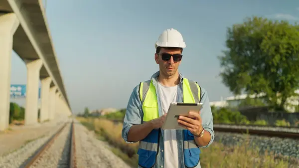 Male foreman engineer wearing helmet and safety vest using tablet working on railway. Engineer checking construction process railway on tablet. Transportation, Engineer leadership concept