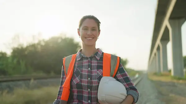 Beautiful engineer woman in safety uniform and helmet standing on railroad smiling and looking at camera. Engineer leadership concept