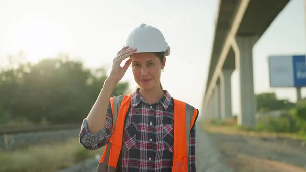 Beautiful engineer woman in safety uniform and helmet standing on railroad smiling and looking at camera. Engineer leadership concept