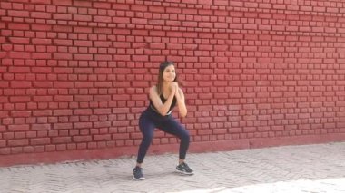 Video of beautiful girl in gym outfit doing squat and knees to chest aerobic exercise standing in front of red brick wall.