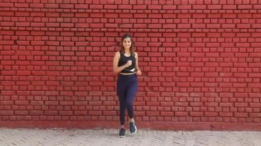 Video footage of Indian woman performing March in place workout standing in front of red brick wall.