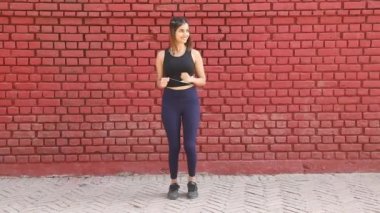Video footage of Indian woman performing March in place workout standing in front of red brick wall.