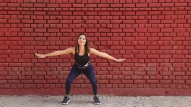 Video of slim woman doing balancing squat exercise standing in front of red brick wall.