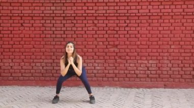 Video of Indian woman in gym suit doing squat and feet together cardio aerobic exercise standing in front of red brick wall.
