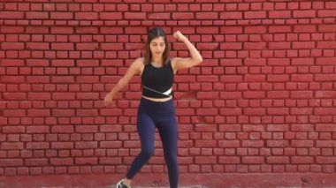 Video of fit girl in gym outfit doing squat and knees to chest aerobic exercise standing in front of red brick wall.