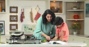 Anne, oğlunun mutfakta yemek pişirirken ders çalışmasına yardım ediyor.