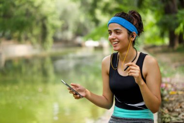 Spor kıyafetli, kulaklıklı, nehir manzaralı sağlıklı bir kadın müzik dinliyor. 