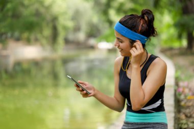 Spor salonundaki kadın parkta beton levhada otururken müzik dinliyor.  
