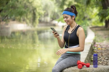 Seksi zinde bir kadın parkta spor yaptıktan sonra dinleniyor ve telefona bakıyor. Sosyal medya paylaşımları okuyor ve spor yaptıktan sonra rahatlıyor.