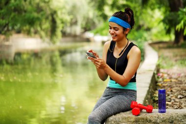 Spor kıyafetli sağlıklı bir bayan parkta egzersiz yaptıktan sonra cep telefonu kullanıyor.