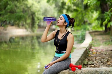 Parkta beton bir levhada otururken şişeden su içen güzel genç bir kadın.