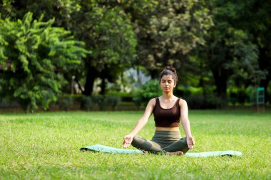 Esmer model meditasyon pozunda yeşil parktaki yoga minderine otur.