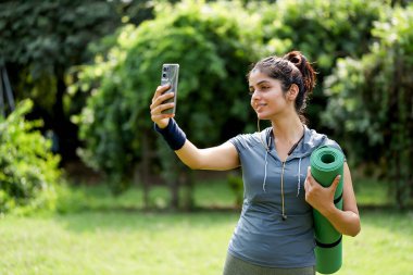 Gülümseyen jimnastik kıyafetli kadın elinde yoga minderiyle parkta selfie çekiyor.