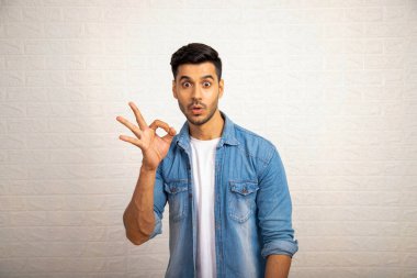 Beyaz tişört ve kot ceket giyen Asyalı adam kameraya bakarken süper iyi işareti gösteriyor.