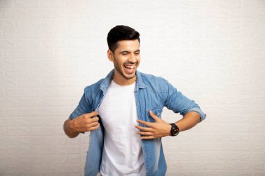 Genç sırıtan adam gülüyor ve gömleğini iki eliyle tutarken beyaz arka planda duruyor.