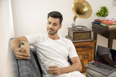 Oturma odasında koltukta oturmuş gülümseyen bir adam cep telefonuyla sosyal medyada makara izlerken telefon kullanıyor.