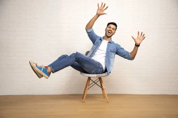 Akıllı görünümlü bir adam, ayakları havada poz veriyor ve beyaz arka planda uzanan kollarıyla sandalyede oturuyor.