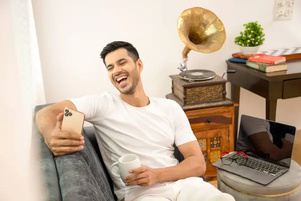 Elinde beyaz kahve fincanı tutan adam cep telefonunda sosyal medyada sırıtıyor.