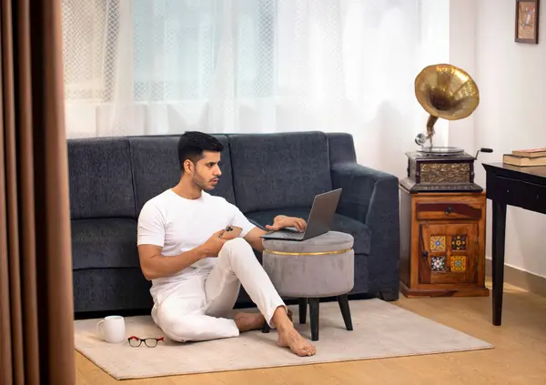 Yakışıklı genç adam elinde cep telefonu tutarken dizüstü bilgisayarda yazıyor.