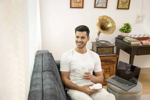 Elinde kahve fincanı ve cep telefonu tutan beyaz kıyafetli seksi Hintli adam koltukta oturup geleceği düşünüyor.