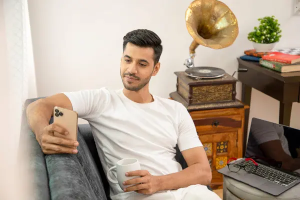 Oturma odasında koltukta oturmuş gülümseyen bir adam cep telefonuyla sosyal medyada makara izlerken telefon kullanıyor.