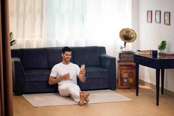 Beyaz tişörtlü, gülümseyen bir adam yerde oturmuş video görüşmesinde konuşuyor ve elinde bir fincan kahve tutuyor.