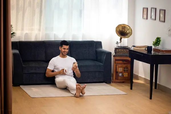 Beyaz tişörtlü gülümseyen genç adam bacak bacak üstüne atarak oturmuş, akıllı telefondan sohbet ediyor ve kahvenin tadını çıkarıyor.