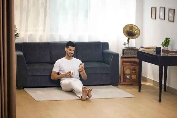 Bacak bacak üstüne atmış beyaz t-shirt giyen cesur bir adam internette cep telefonuyla alışveriş yapıyor ve elinde kahve tutuyor.