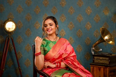 Hintli sari giyen sevimli bir kadın model kameranın önünde oturur ve bir sandalyede poz verirken gülümser.