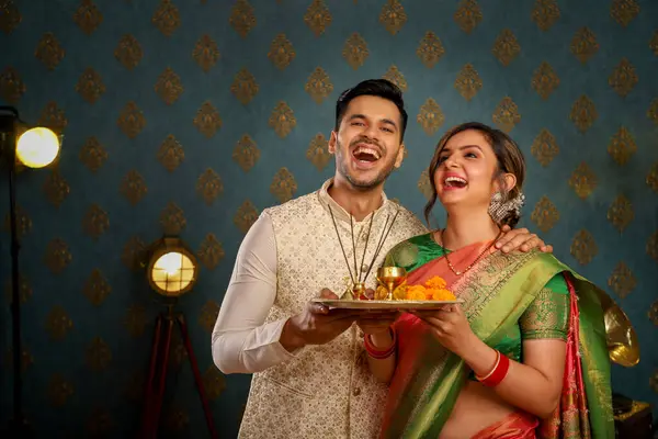 Geleneksel Hint kıyafetleri içinde ellerinde puja tabağı ve gülümseyen bir Diwali çifti resmi.