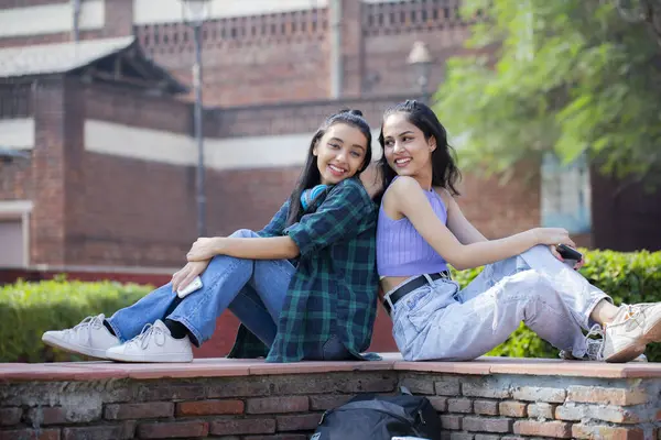 Fashionable females are smiling and sitting back-to-back in the college park