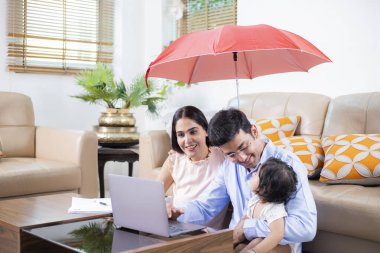 Family Insurance concept where mother, father and baby under umbrella while sitting at home clipart