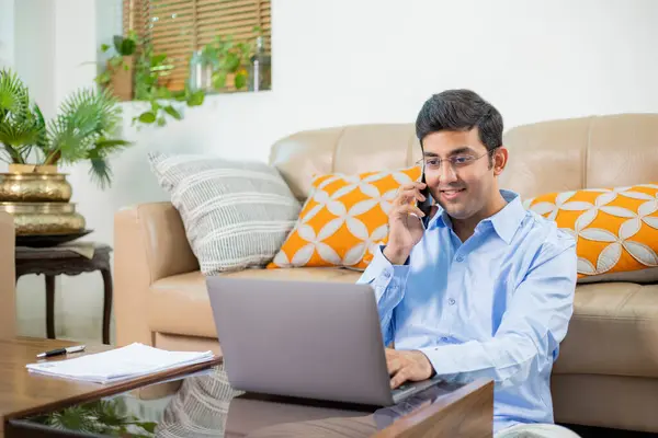 Mavi gömlekli mutlu Hintli adam dizüstü bilgisayarla çalışırken cep telefonuyla konuşuyor.