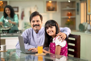 Yakışıklı baba ve kız kamera önünde poz verirken anne arka planda çalışıyor.