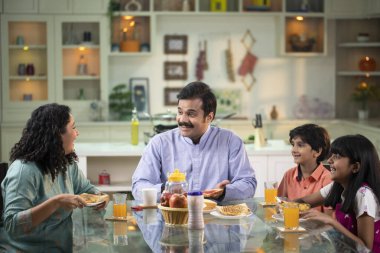 Aile mutfakta kahvaltı yapıyor, sohbet ediyor ve gülüyor.