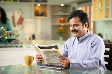 Adam evde yemek masasında otururken gazete okuyor.