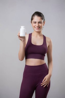 Mor spor kıyafetleri içinde spor salonu modeli kameranın önünde duruyor, elinde bir takviye şişesi var.