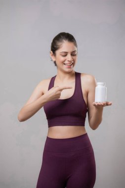 Bir sağlık modeli gülümsüyor, ayakta duruyor ve parmağını tablet şişesine doğrultuyor.