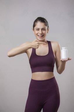 Spor kıyafetleri içinde güzel bir manken gülümser ve parmağını tablet şişesine doğrultur.